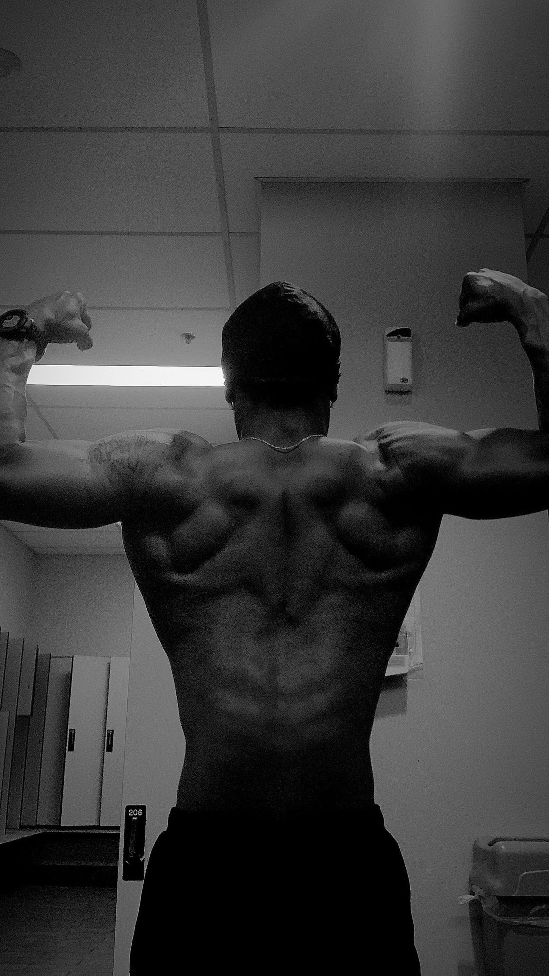 Black and white photo of a muscular individual from the back, showcasing a well-defined upper body and broad shoulders in a gym setting with visible equipment in the background.